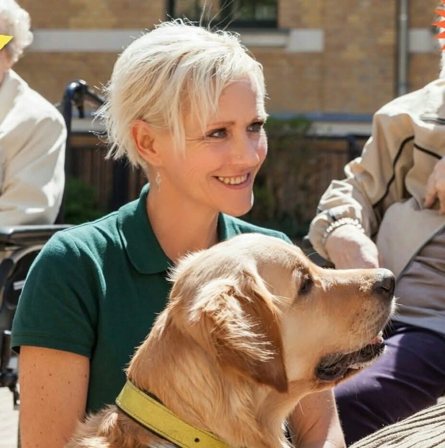 Eine blonde Frau mit einem Golden Retriever, umgeben von älteren Personen im Hintergrund.